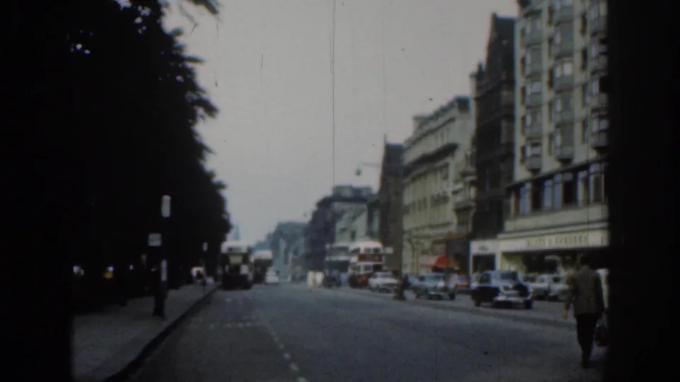 Final 1961 London street frame showing cars, buses, shopfronts, a dark tree line, and a pedestrian walking near the right edge.