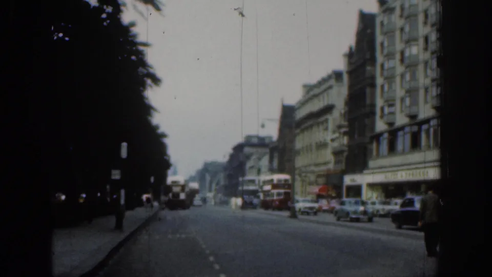 1961 London street view with double-decker buses, period cars, pedestrians, and tall buildings along a tree-lined avenue.