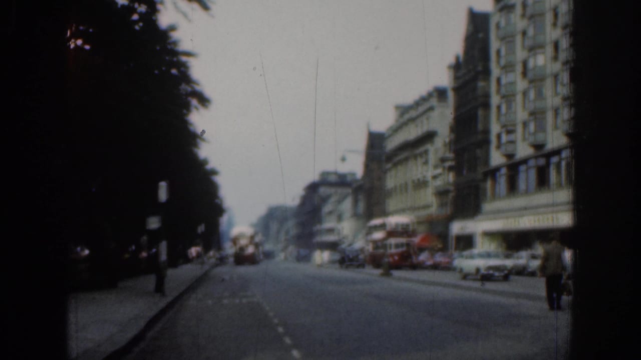 London avenue scene with red buses, cars, pedestrians, storefront buildings, and dark trees framing the road.