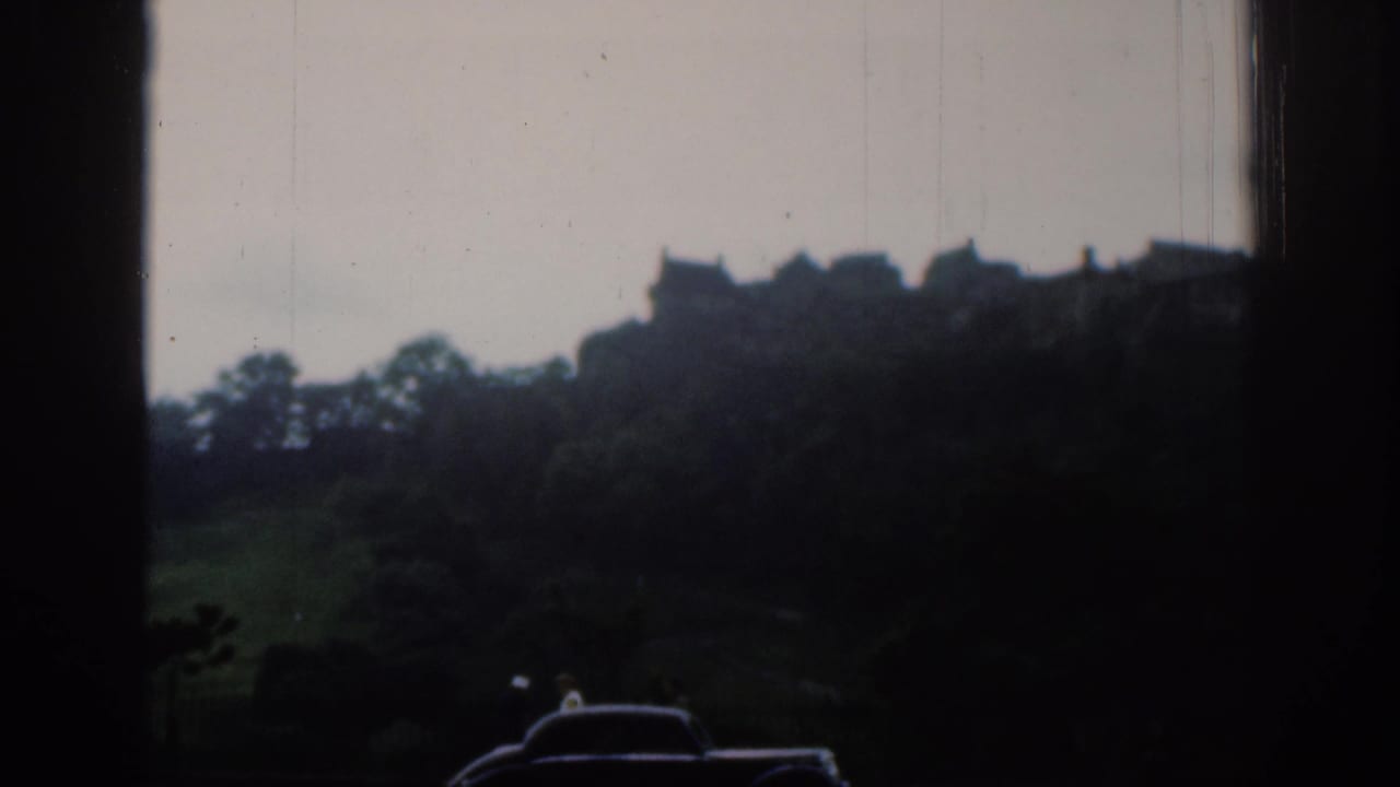 Soft 8mm view of hilltop buildings and a castle-like silhouette above trees, with cars passing in the foreground.