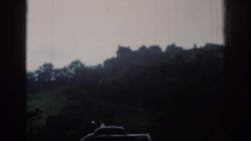 Soft 8mm view of hilltop buildings and a castle-like silhouette above trees, with cars passing in the foreground.