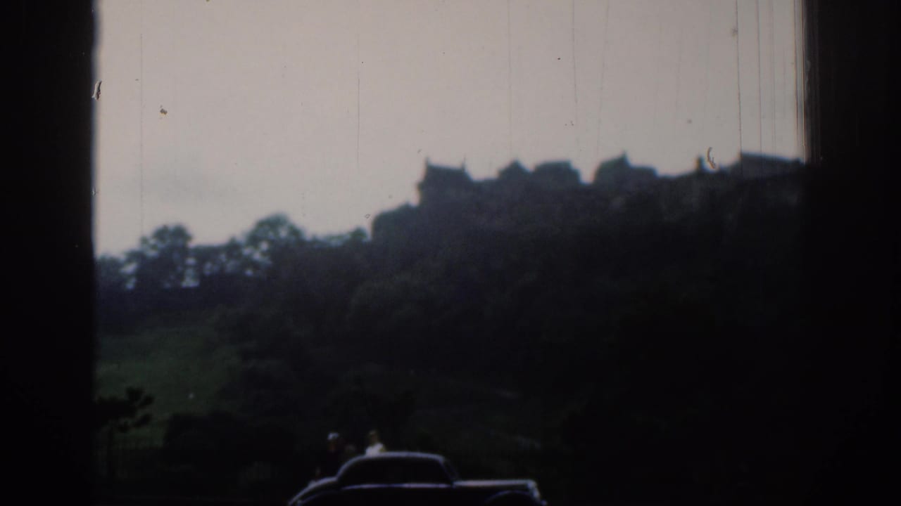 1961 home-movie view toward the metadata-identified Edinburgh Castle, framed by a dark window edge with a car and distant people below.
