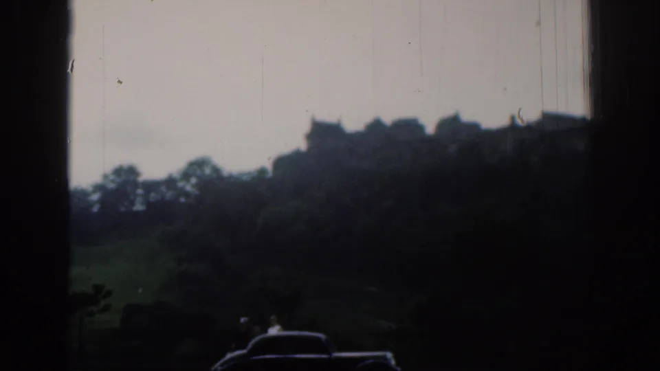 1961 home-movie view toward the metadata-identified Edinburgh Castle, framed by a dark window edge with a car and distant people below.