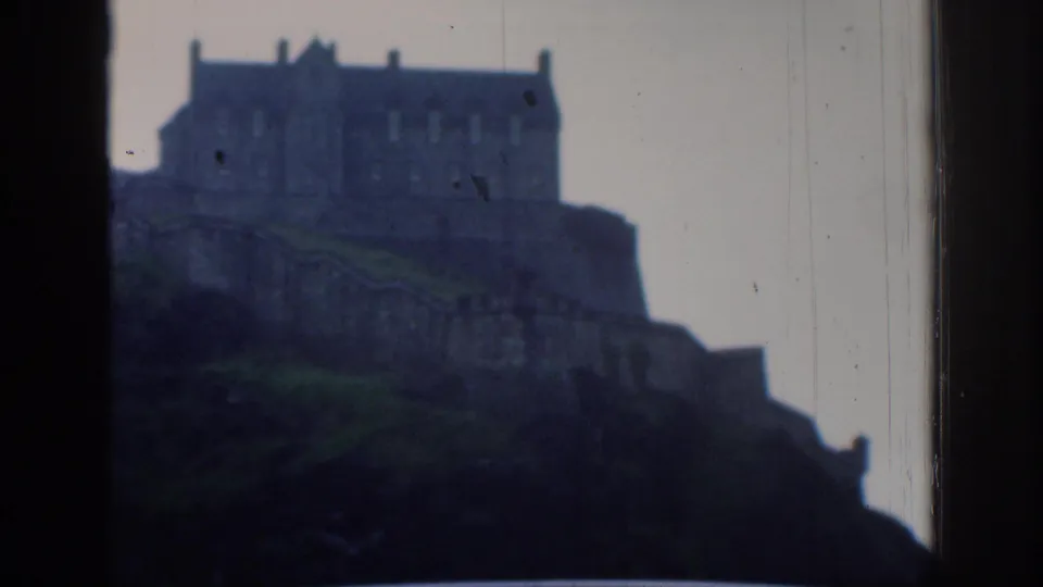 Soft, scratched 8mm frame of the metadata-identified Edinburgh Castle on its rocky rise, viewed from a roadside or windowed position.