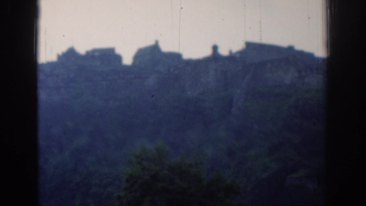 Distant buildings silhouetted above a dark wooded hillside, framed by black window edges.