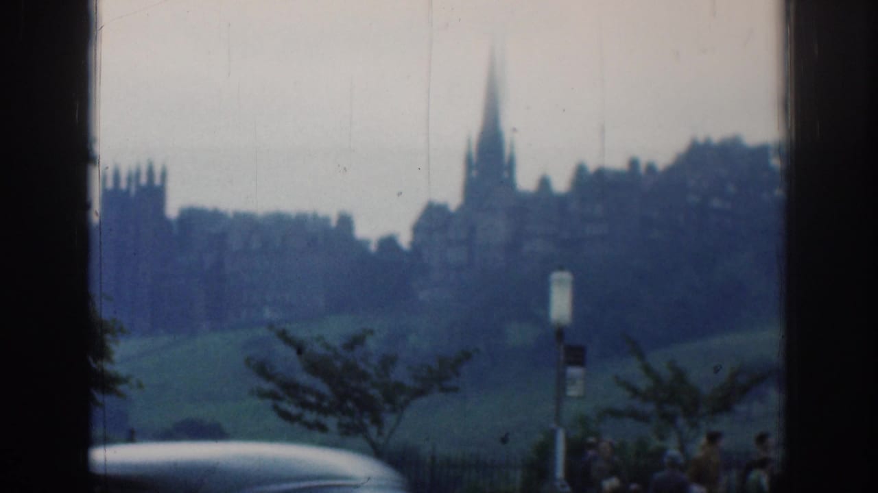 Distant church spire and buildings beyond a green slope, seen through dark vertical window frames.