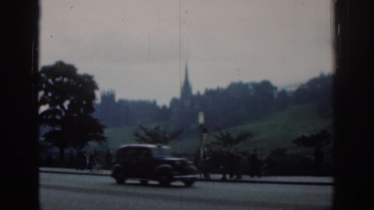 A dark car travels along a London road in 1961, with distant Gothic-style architecture, trees and small roadside figures beyond it.