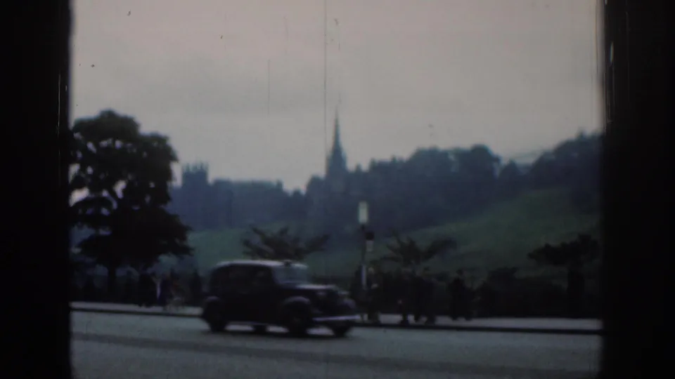 A dark car travels along a London road in 1961, with distant Gothic-style architecture, trees and small roadside figures beyond it.