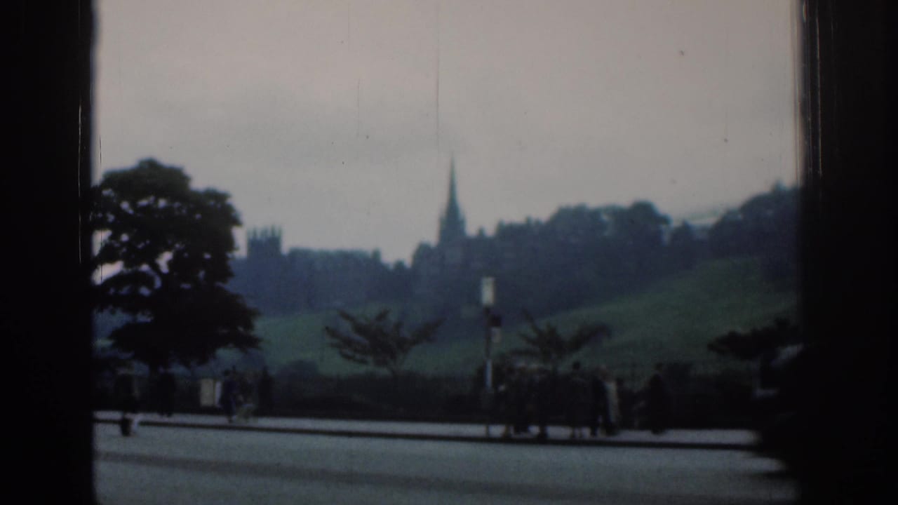 1961 London 8mm frame showing a distant spire on a green rise, trees, roadside pavement, and small pedestrian figures.