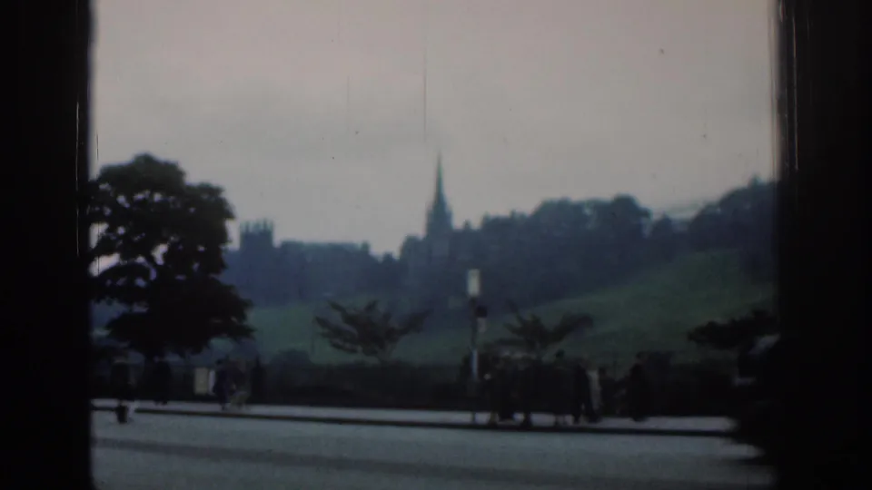1961 London 8mm frame showing a distant spire on a green rise, trees, roadside pavement, and small pedestrian figures.