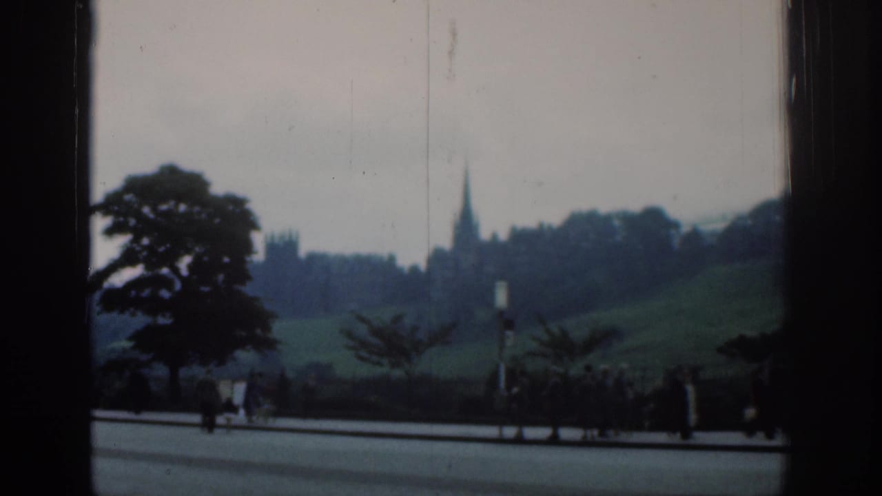 1961 London 8mm frame with pedestrians near a roadside and a distant spire partially framed by dark architectural edges.
