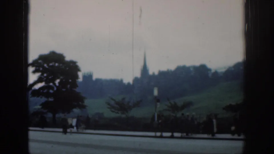 1961 London 8mm frame with pedestrians near a roadside and a distant spire partially framed by dark architectural edges.