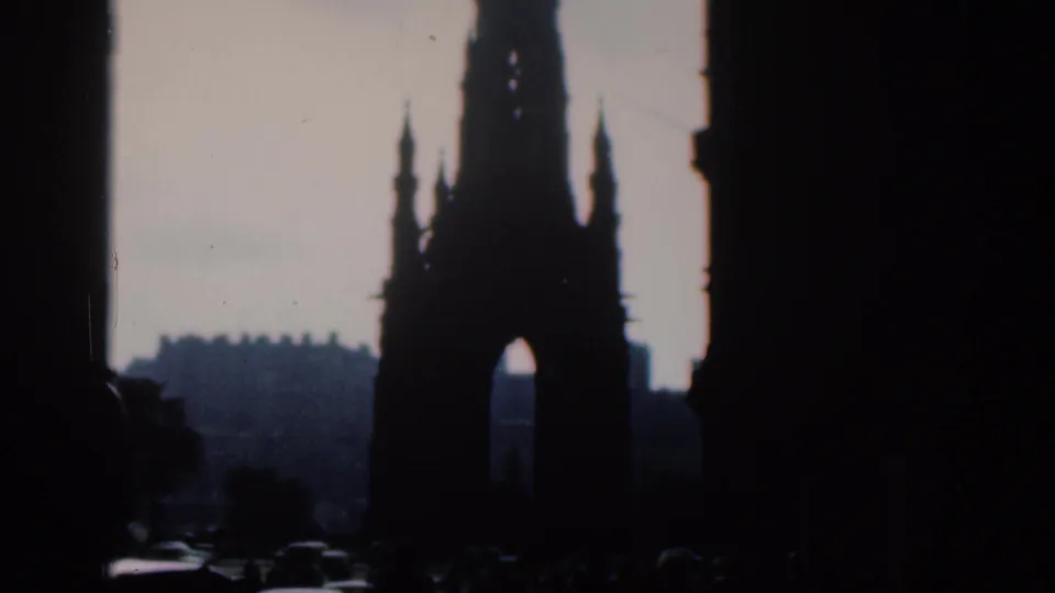 A silhouetted Gothic monument with an arched opening stands beyond shadowed street architecture in the 1961 home-movie frame.