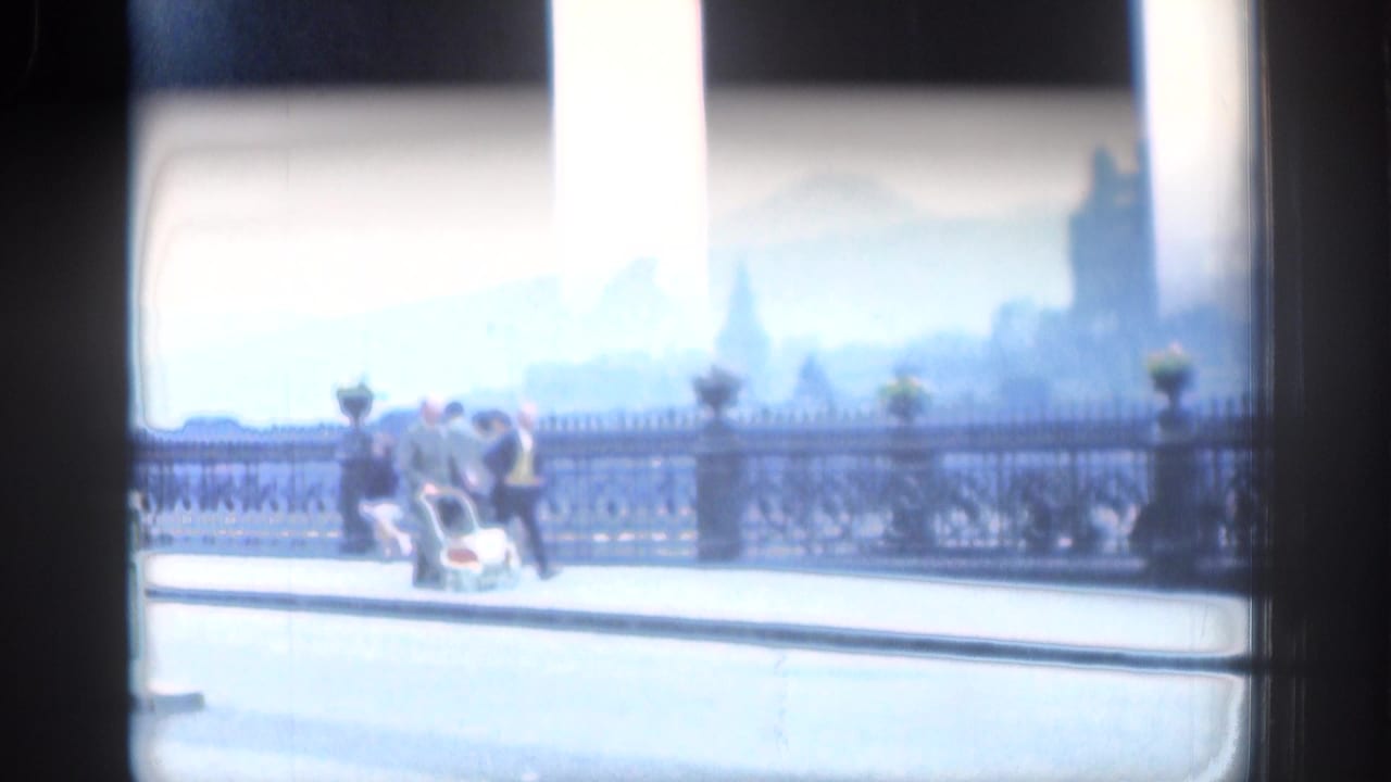 Pedestrians and a baby carriage seen beyond dark window framing beside ornate railings.