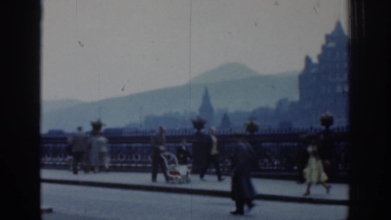 1961 London street view with adults walking near ornate railings, a pram, distant buildings, and a hazy hill beyond.