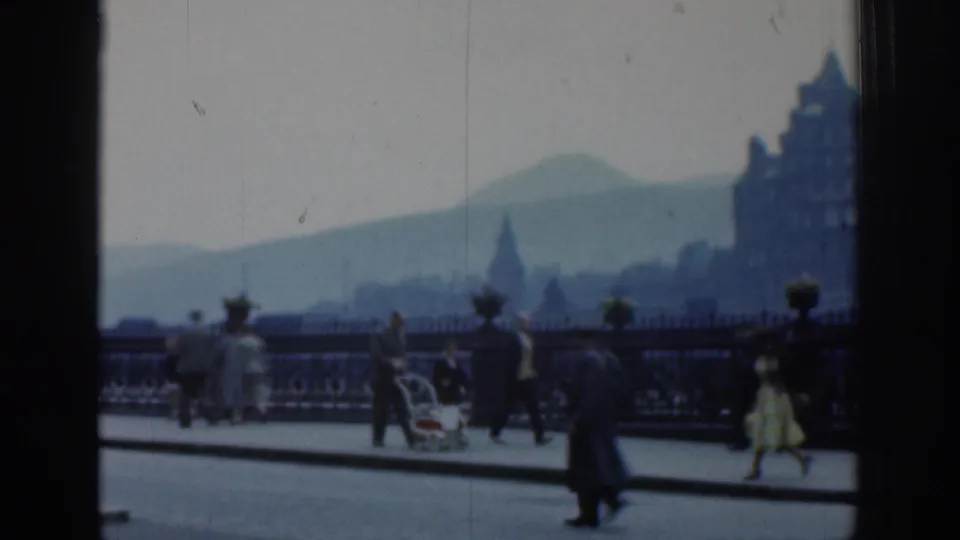 1961 London street view with adults walking near ornate railings, a pram, distant buildings, and a hazy hill beyond.