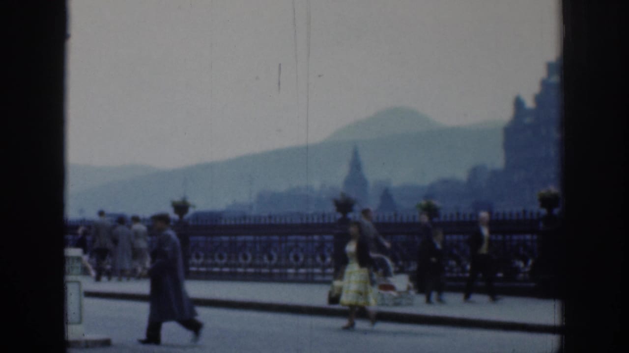 1961 London home-movie frame with a woman in a yellow patterned dress and other pedestrians beside ornate railings.