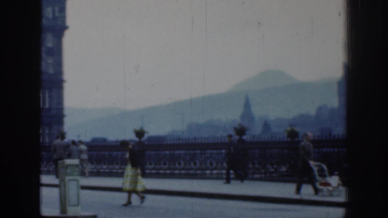 1961 London street scene showing pedestrians, a baby carriage, ornate railings, and a hazy distant hill through home-movie softness.