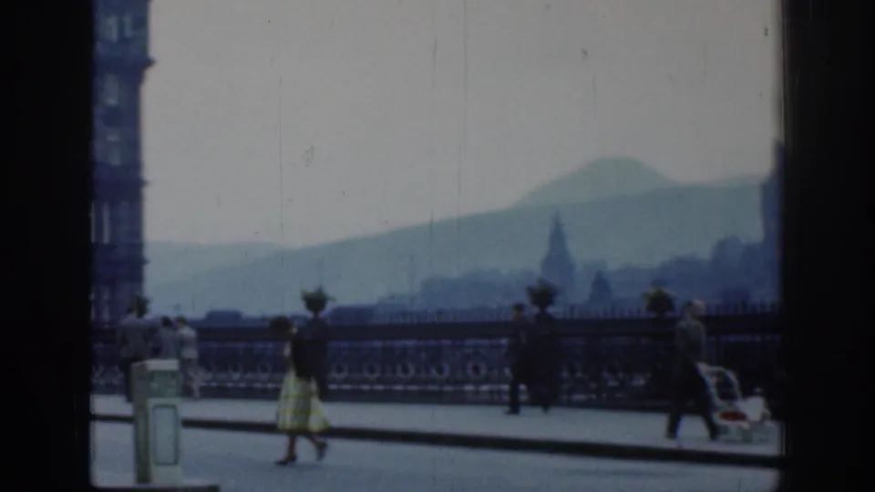 1961 London street scene showing pedestrians, a baby carriage, ornate railings, and a hazy distant hill through home-movie softness.