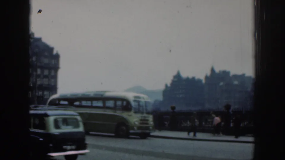 1961 London street view with a green-and-cream bus, a small car, pedestrians, railings and historic-looking buildings in the background.