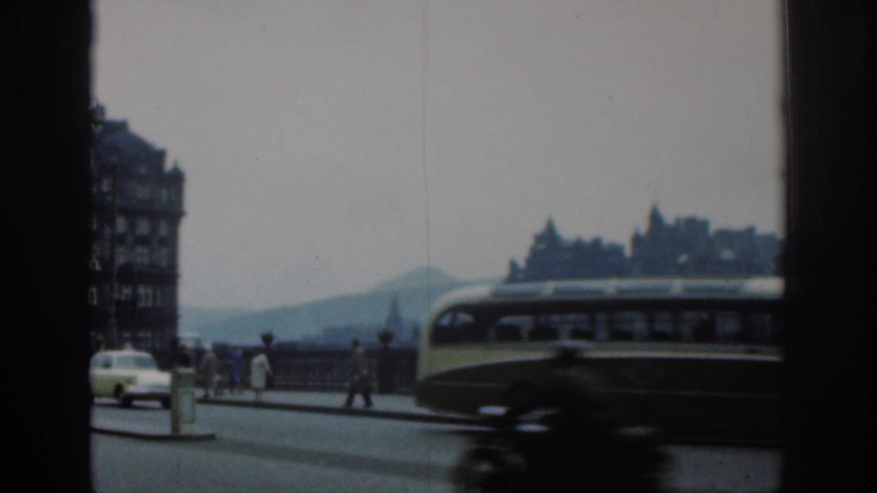 1961 London home-movie frame with a bus crossing the foreground, pedestrians along the railing and distant buildings under a pale sky.