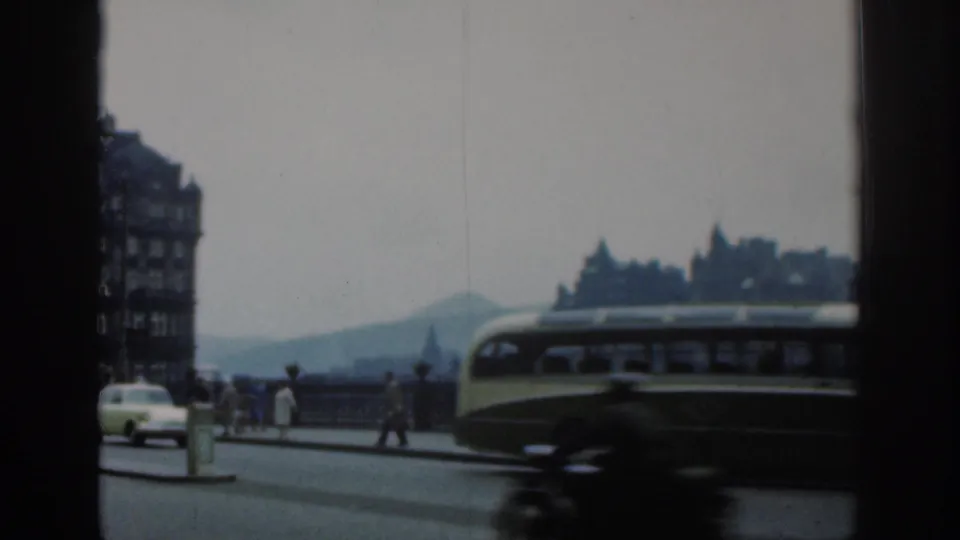 1961 London home-movie frame with a bus crossing the foreground, pedestrians along the railing and distant buildings under a pale sky.