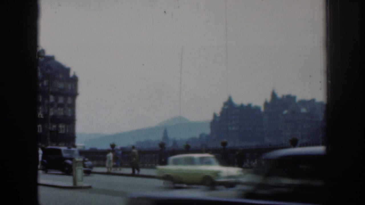 1961 London street frame showing classic cars moving past bridge railings, pedestrians, hazy buildings and a distant landform.