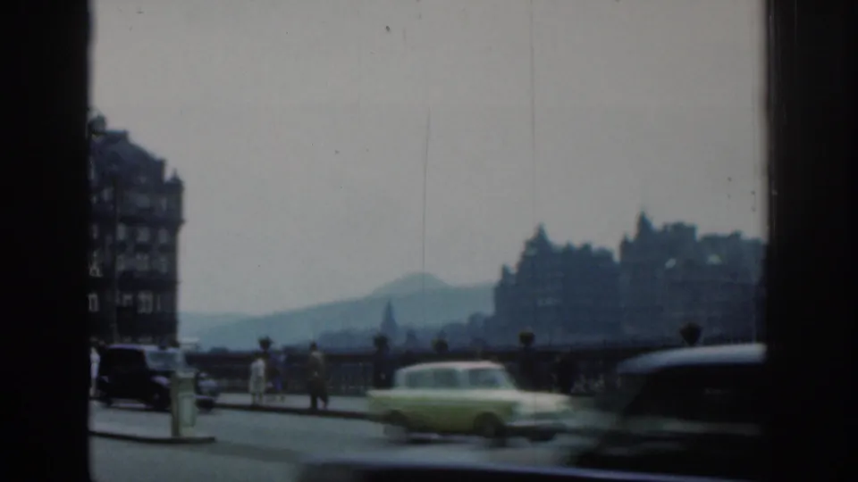 1961 London street frame showing classic cars moving past bridge railings, pedestrians, hazy buildings and a distant landform.