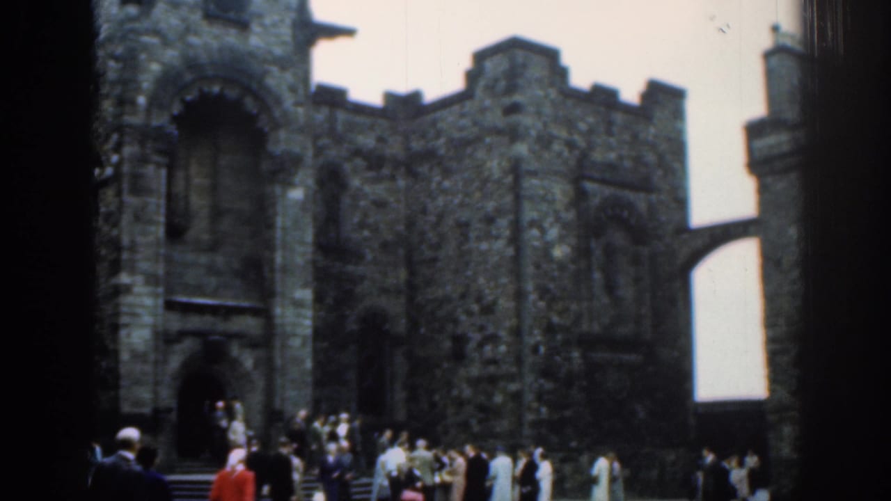1961 London frame showing a stone castle entrance, adjoining crenellated wall, bridge-like passage, and tourists in front.