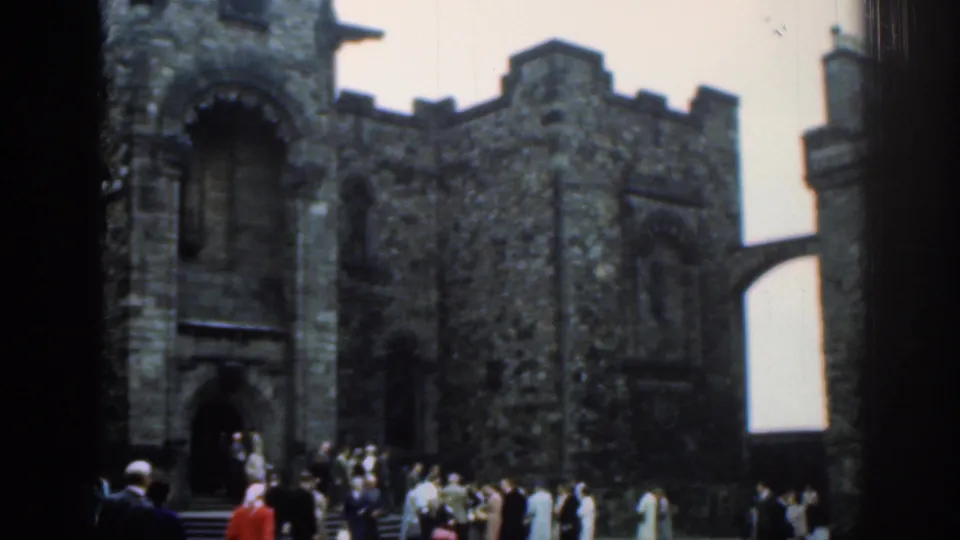 1961 London frame showing a stone castle entrance, adjoining crenellated wall, bridge-like passage, and tourists in front.
