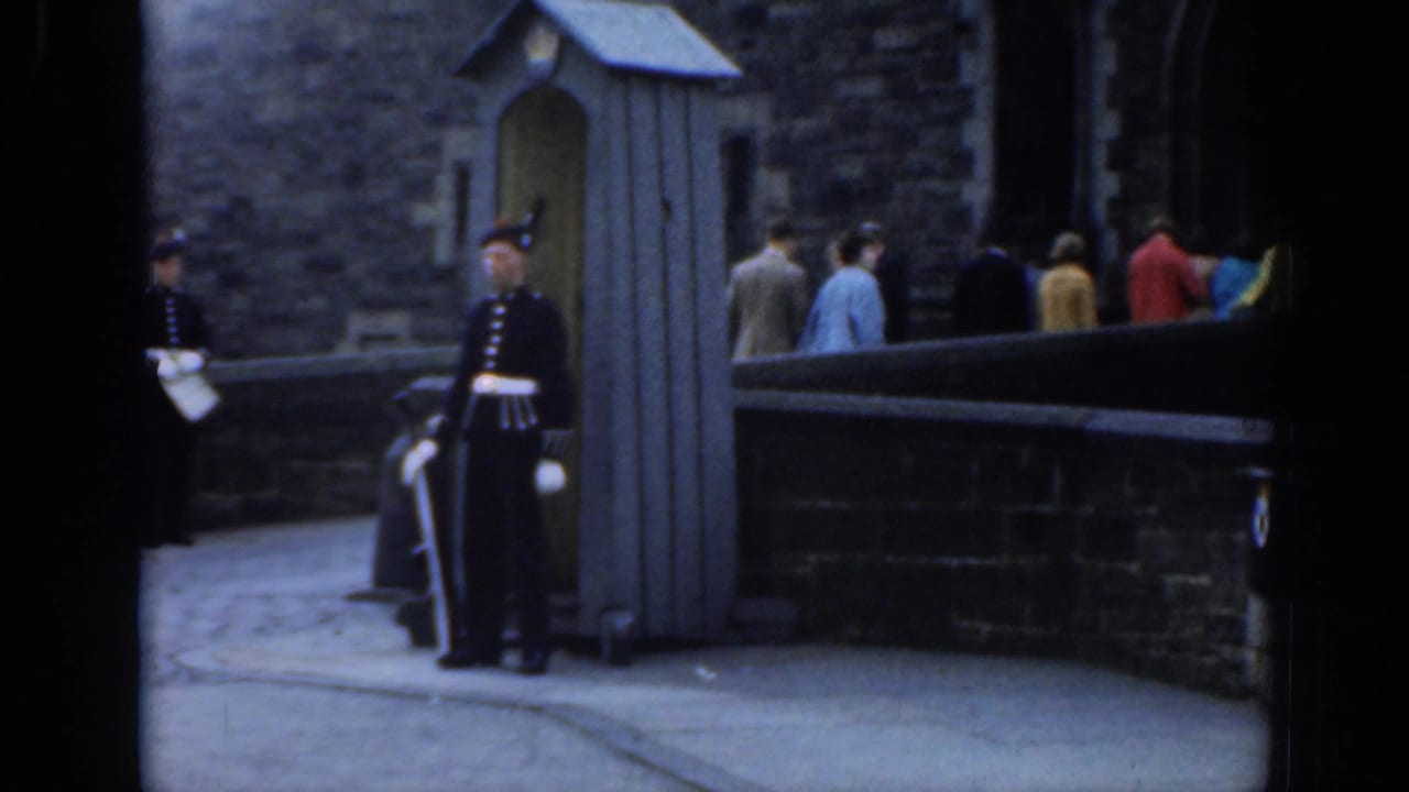 A uniformed guard in London stands beside a small sentry box as pedestrians pass along a stone-walled public viewpoint.