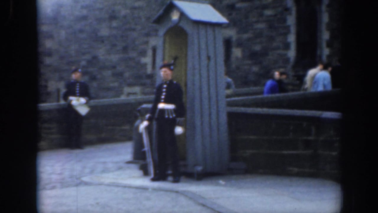 1961 London frame with guards in dark uniforms and tall hats standing beside a sentry box as tourists pass near a historic stone exterior.