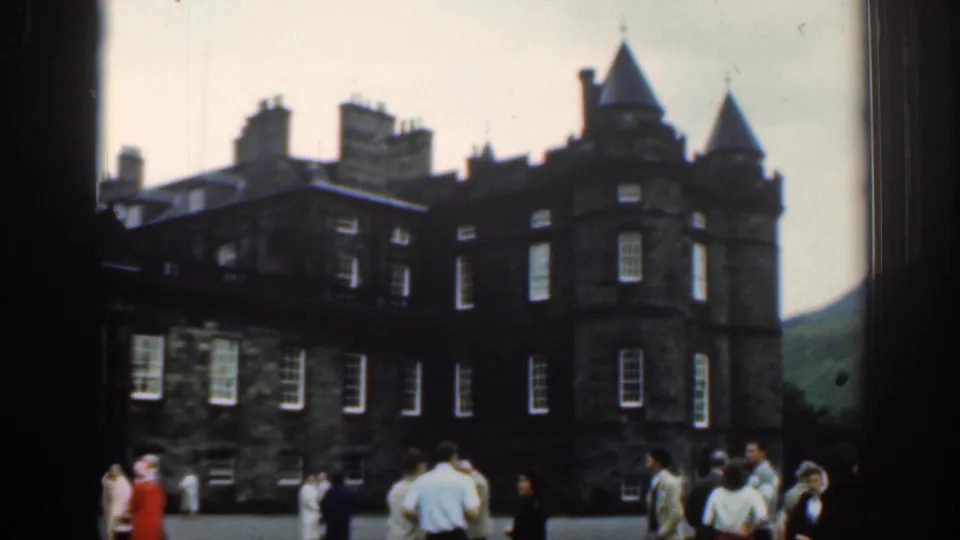 1961 travel frame showing a large dark historic building with turrets, chimneys, windows, and tourists gathered in the foreground.