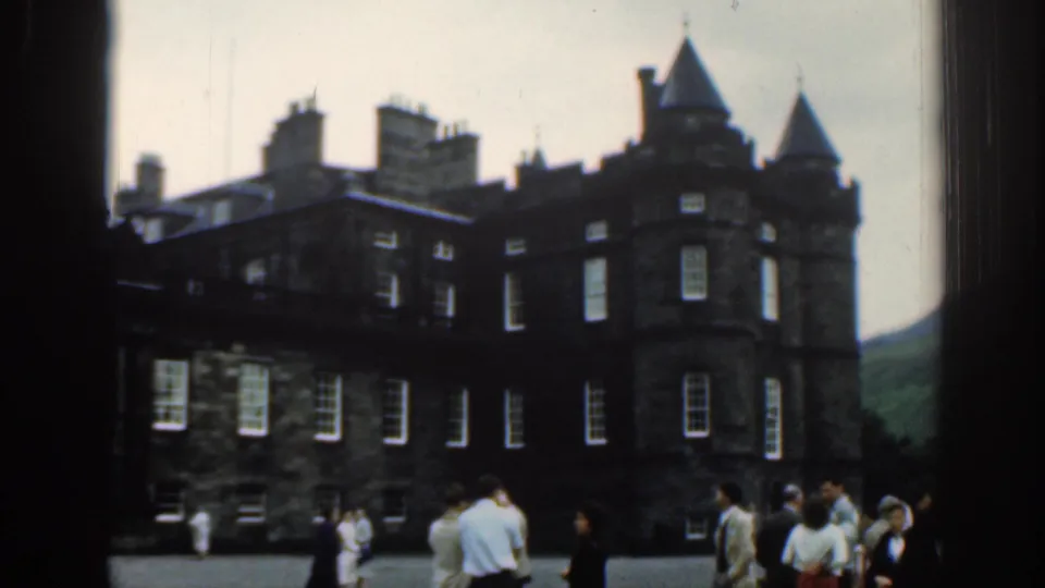 1961 London home-movie frame showing visitors gathered near a turreted stone historic building under cloudy daylight.