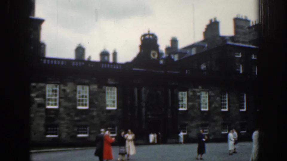 1961 London visitor footage with people spaced across a courtyard before a historic stone building and central clock feature.
