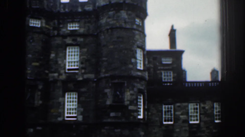 1961 8mm view listed as London, showing a historic dark stone building with rounded towers and white-paned windows through a window frame.