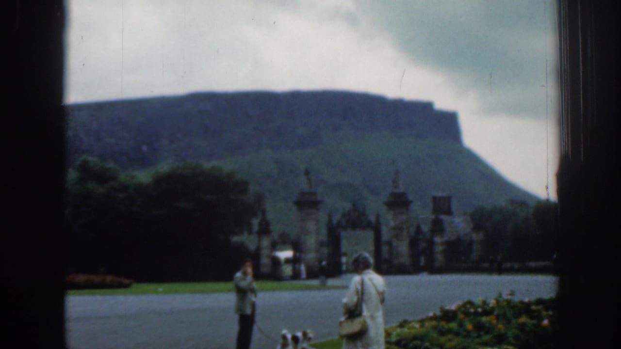 Wide 1961 8mm view of estate grounds with ornate gates, visitors in period clothing, a small dog, flower beds, and a hill beyond.
