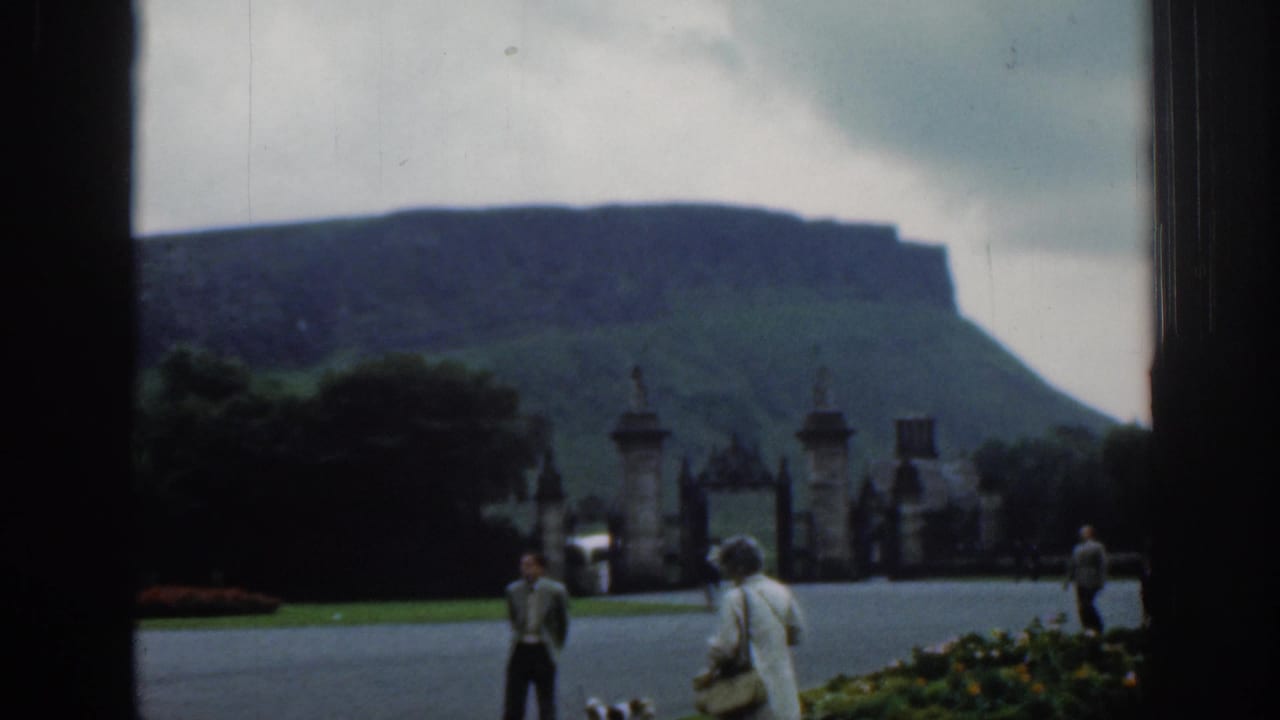 1961 8mm estate-ground view listed as London, with visitors walking near gates, a flower bed, trees, and a broad hill beneath cloudy sky.
