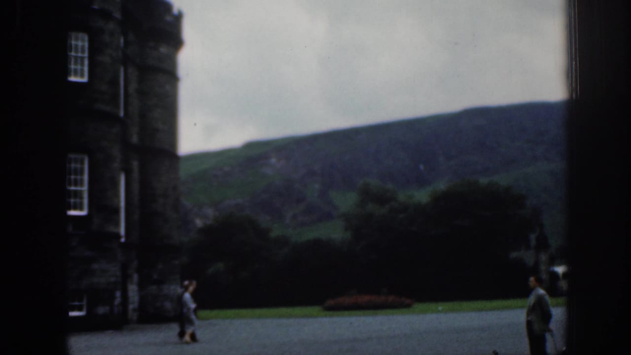 1961 London home-movie frame showing people crossing a courtyard near a tall dark stone building, with green hillside visible in the distance.