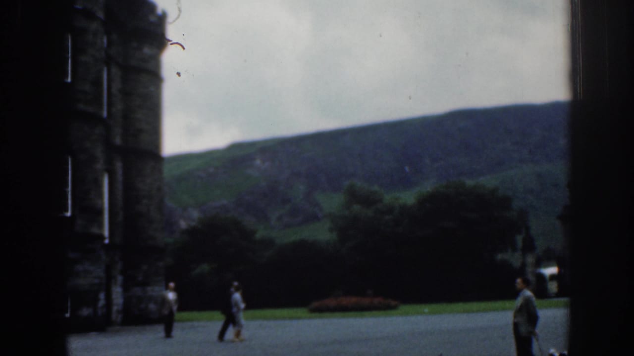 1961 London home-movie view of visitors crossing open paving beside a stone tower, with lawn, trees, and hillside in the background.