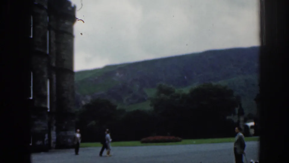 1961 London home-movie view of visitors crossing open paving beside a stone tower, with lawn, trees, and hillside in the background.