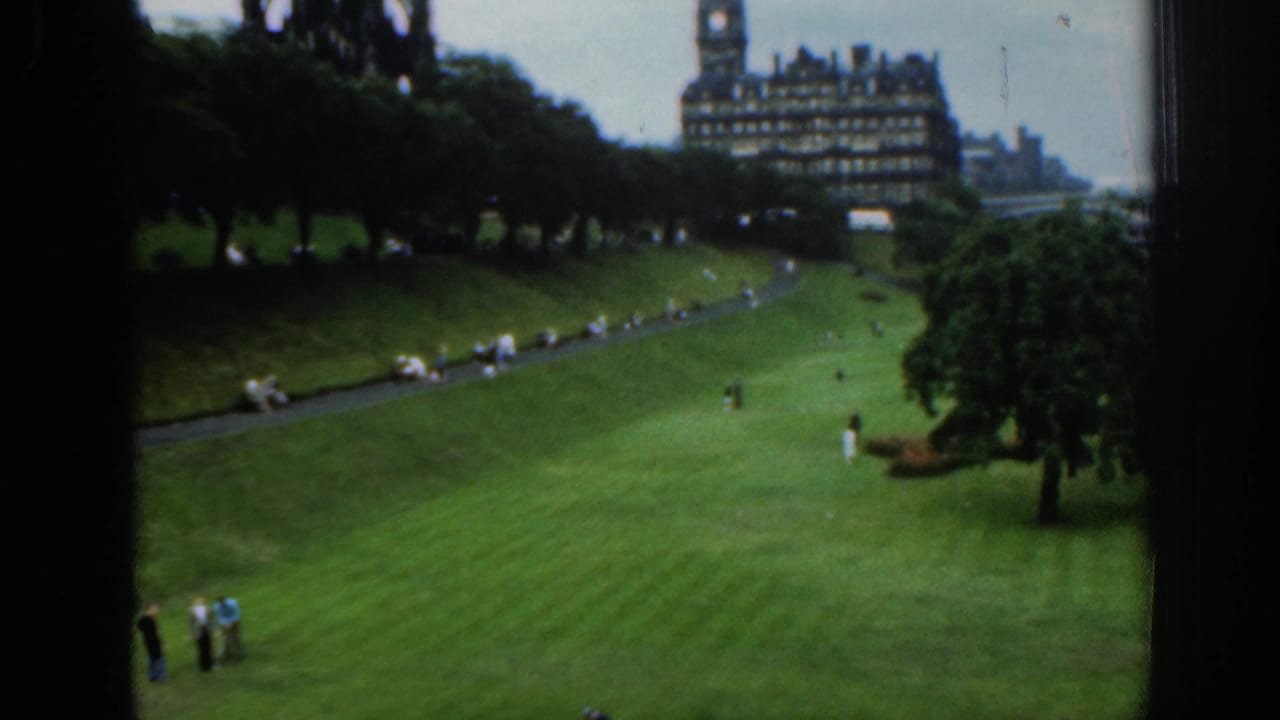 1961 London home-movie frame looking over green lawns, paths, trees, and scattered visitors toward a large ornate building.