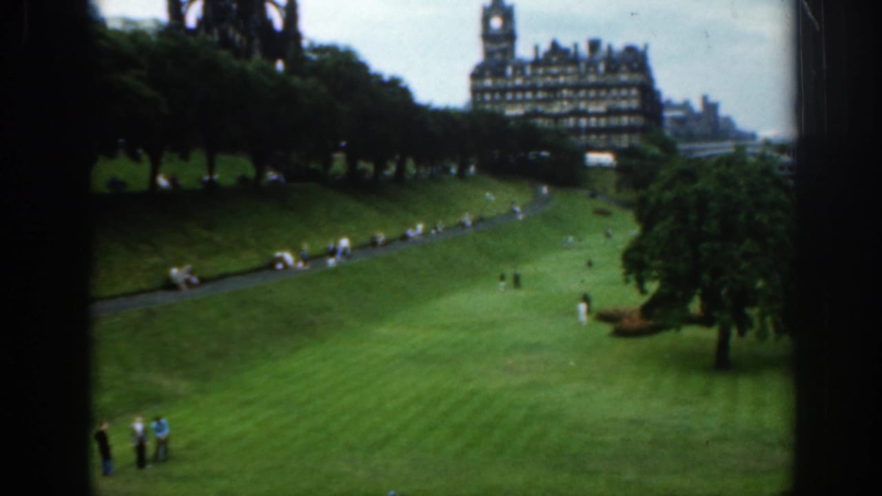 Wide park lawn with people on grass, a curving path, trees, and a distant historic building.