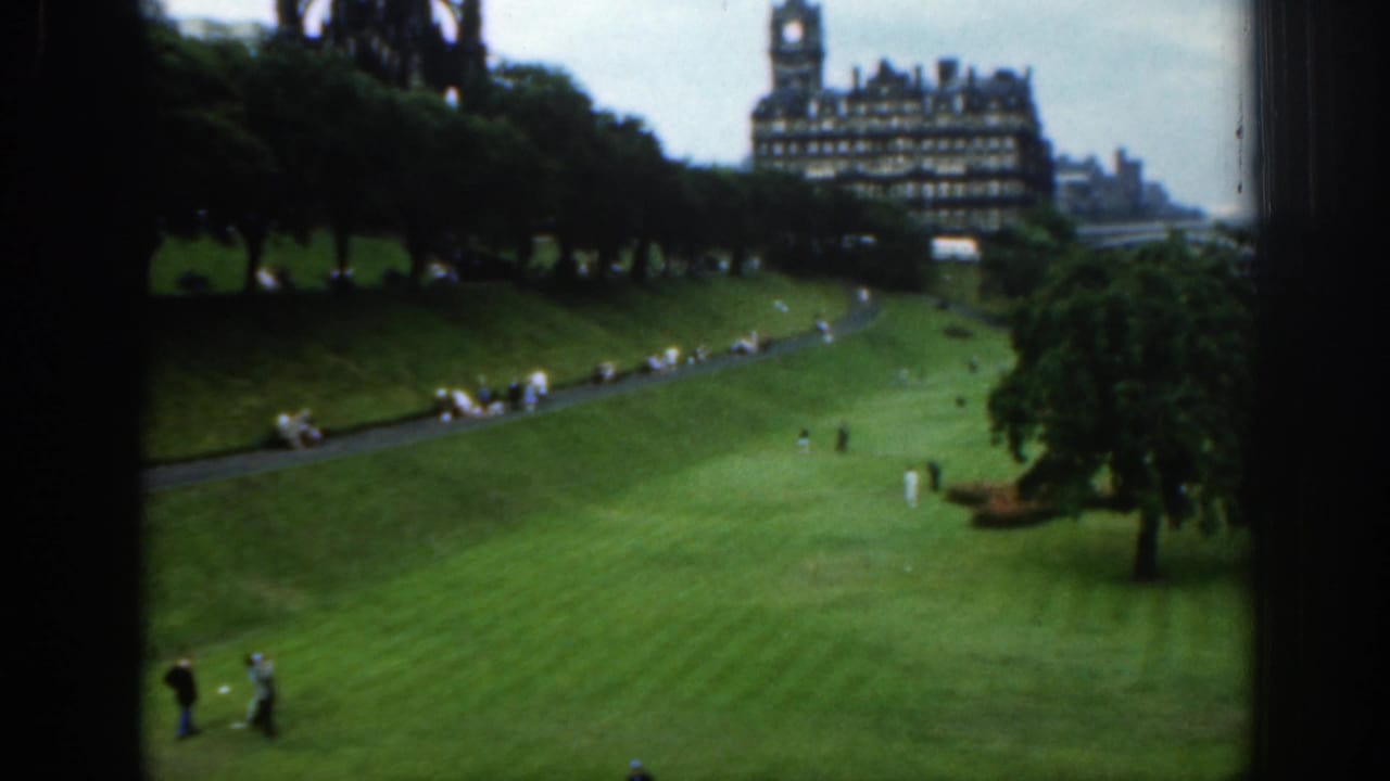 Park lawn and path with small groups of people, trees, and a large old building in the distance.