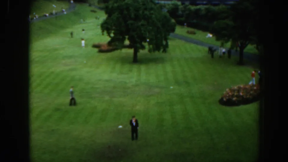 1961 Scotland 8mm frame showing men on a manicured green, with trees, paths, and a flower bed in the background.