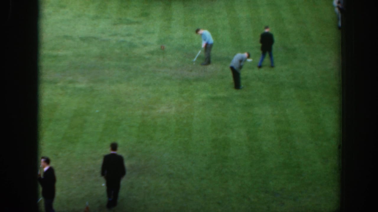 Several men stand and bend over on a green while practicing with golf clubs.