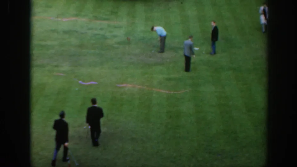1961 Scotland 8mm frame showing men in coats and casual shirts gathered around a golf practice area on grass.
