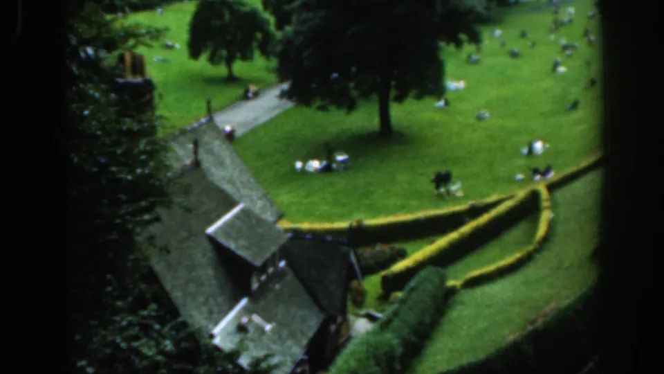1961 Scotland park view showing people gathered on the grass beside a curving path, hedges, trees, and a small roofed structure.