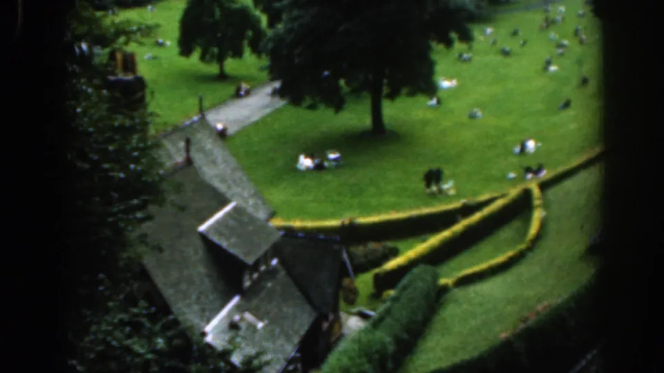 1961 Scotland 8mm frame looking down on a public park with seated groups, a winding path, hedges, mature trees, and a small structure.