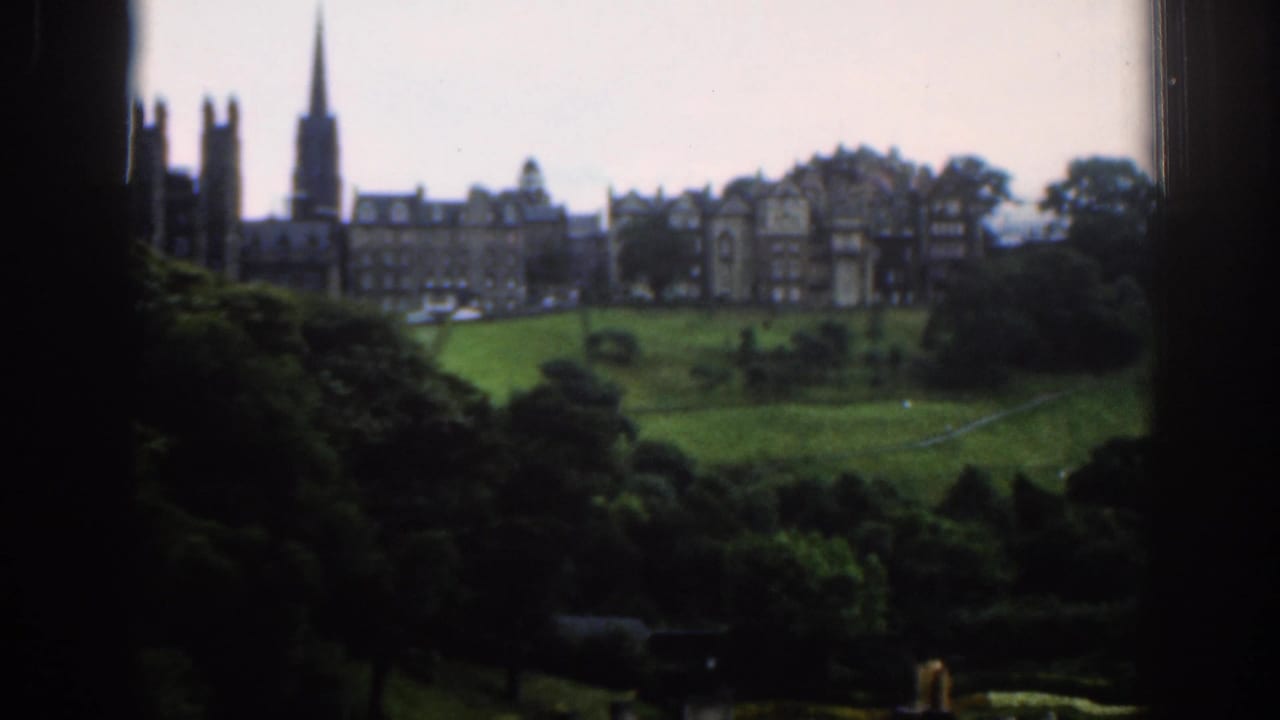 Distant stone buildings and spire above a green hillside and trees.